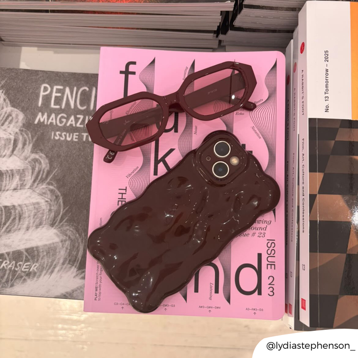 Burgundy 3D bubble phone case on a pink magazine, beside pink glasses and stacked books.