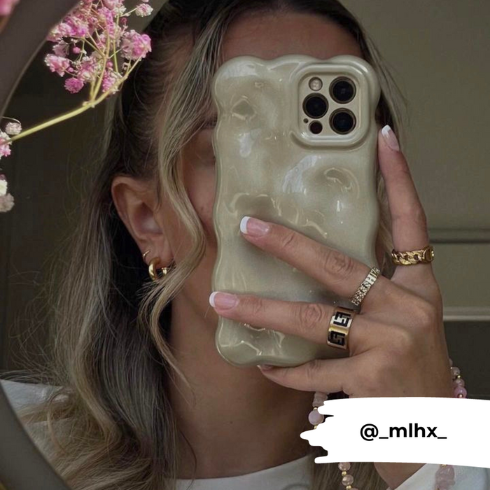 A person with wavy hair holds an iPhone 15 featuring the Coconut Lane 3D Pearl Phone Case - Gold in front of their face. They wear gold hoop earrings, a gold ring, and have manicured nails. There are pink flowers visible in the upper left corner. The Instagram handle @_mlhx_ is displayed at the bottom.