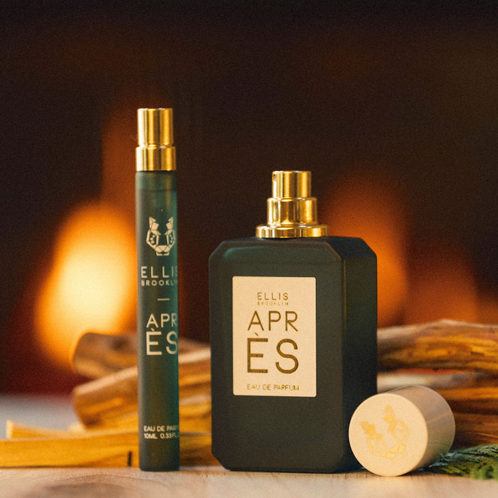 Apres 10ml Eau De Parfum Travel Spray next to Apres 50ml Eau De Parfum with cap removed on wooden table with pieces of wood and small green leaves, with fire in background.