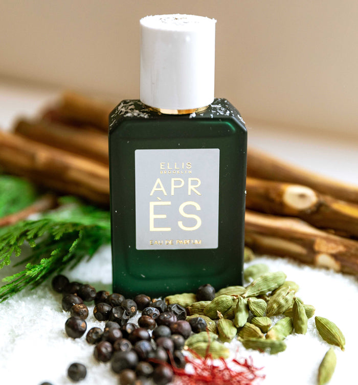 Apres 50ml Eau De Parfum with snow lightly covering bottle on snow with berries, saffron, green leaves, and pieces of wood, against tan background.