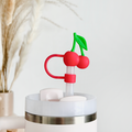 The Cherries Straw Cover by Coconut Lane features a white tumbler adorned with a silver rim, complemented by a uniquely designed silicone straw topped with a red cherry-shaped decoration and green leaves for added durability. The cover ensures the straw remains hygienic, while in the background, a vase filled with dried pampas grass enhances the minimalist and clean aesthetic.