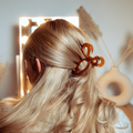 Woman with long blonde hair styled in a brown bow claw clip, standing in front of a lit vanity mirror.