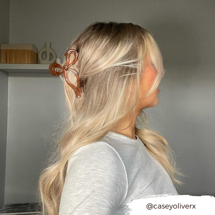 Woman with long wavy blonde hair wears a brown bow claw clip, facing sideways in a modern room.