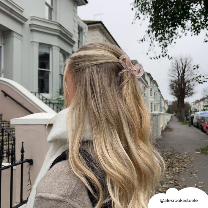 Woman with long blonde hair wears a pink bow claw clip while walking down a tree-lined street.