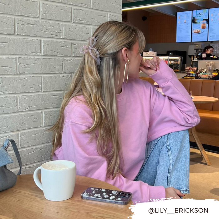 Blonde woman at café wearing a pink bow claw clip, with coffee and phone on the table.