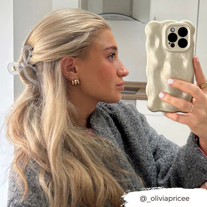 Woman with long blonde hair wearing a white bow claw clip, taking a mirror selfie. Instagram handle is visible.