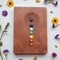 Brown leather notebook with chakra stones and mandala design on a white background with flowers.