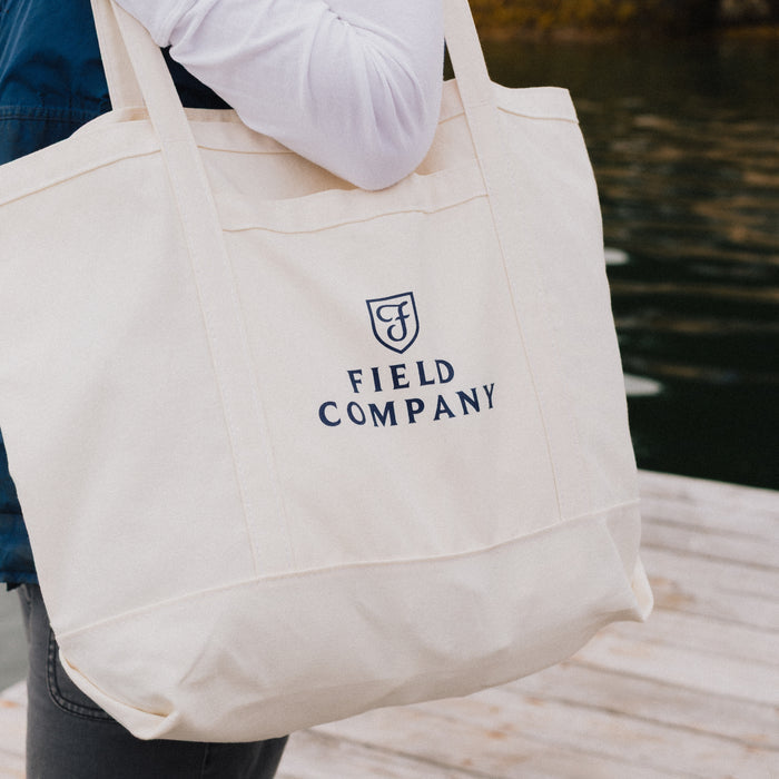 Field Market Tote in natural canvas with navy logo, carried over the shoulder on a waterside dock