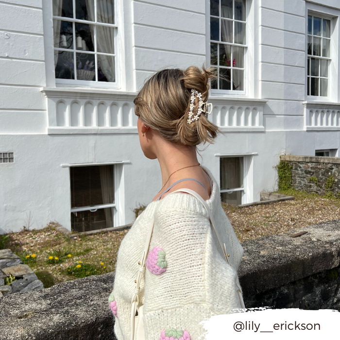 Blonde woman with hair in a bun, wearing a Coconut Lane floral and pearl claw clip, stands by a white building.