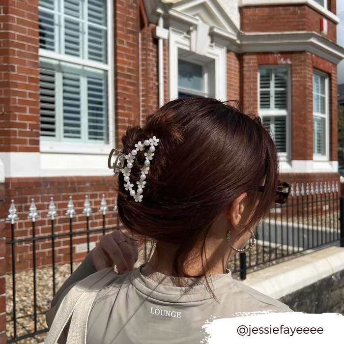 Woman with brown hair wears a floral claw clip with pearls, sunglasses, and hoop earrings beside a brick building.