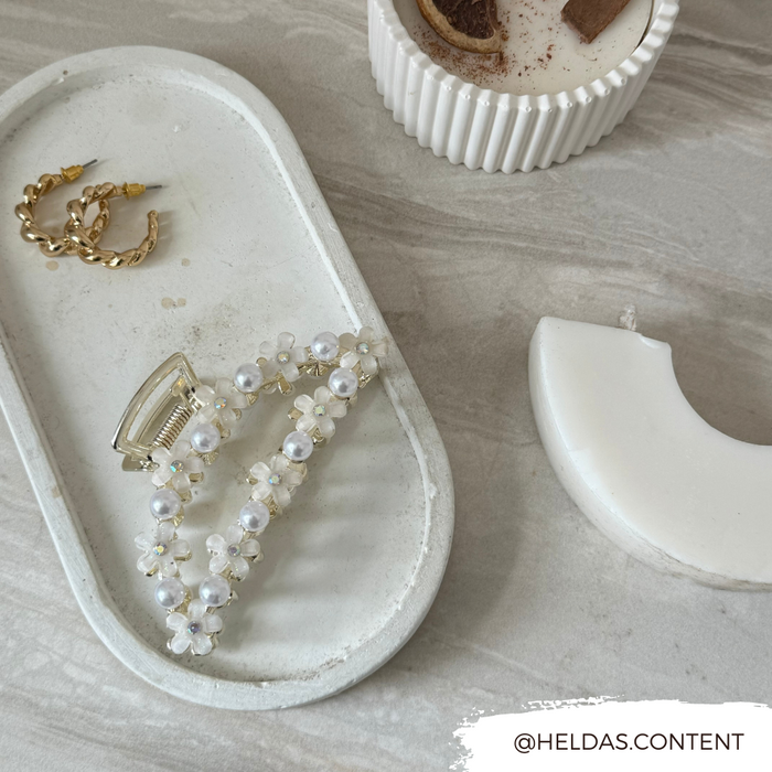 White claw clip with floral and pearl accents, gold hoop earrings, candle, and crescent tray on a light stone surface.