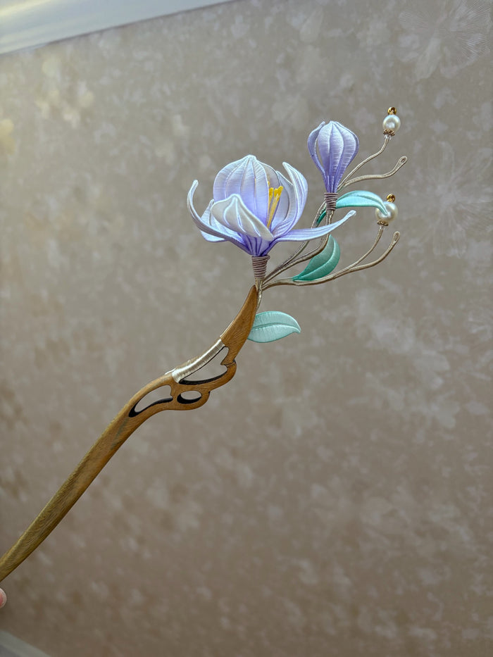 A handmade Chanhua magnolia hairpin with purple flowers and green leaves on a wooden stem.