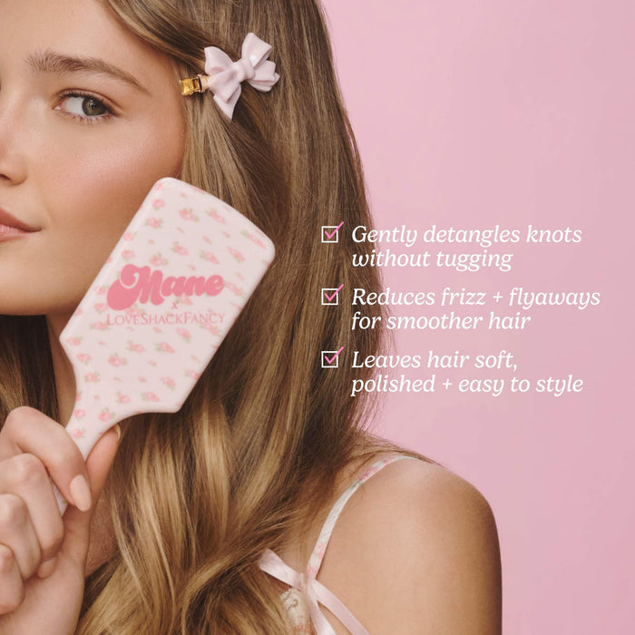 A woman with long light brown hair holds a pink hairbrush with a floral pattern and the brand name 'Mae' printed on it. The background is a solid light pink, with text listing the benefits of the haircare product.