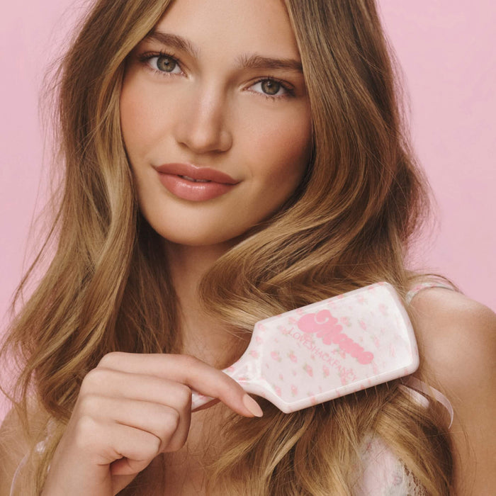A woman with long wavy light brown hair holds a pink hairbrush with a white handle and pink floral pattern against a solid pink background.