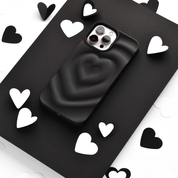 A smartphone encased in Coconut Lane's Melting Heart Phone Case - Matte Black sits on a black surface, surrounded by scattered black and white heart-shaped cutouts. The image emphasizes a monochromatic theme with a focus on the phone and the heart shapes.