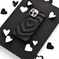 A smartphone encased in Coconut Lane's Melting Heart Phone Case - Matte Black sits on a black surface, surrounded by scattered black and white heart-shaped cutouts. The image emphasizes a monochromatic theme with a focus on the phone and the heart shapes.