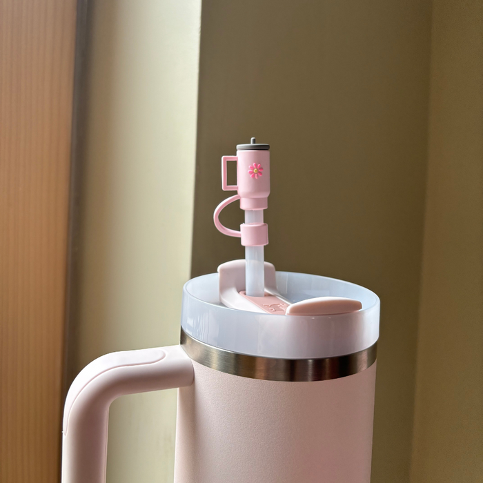 A close-up of Coconut Lane's Mini Pink Tumbler Straw Cover, featuring a silicone spout button for controlling the flow of liquid. The tumbler is set against a beige background, with ambient light adding a warm tone to the scene, and includes a charming matching straw topper.