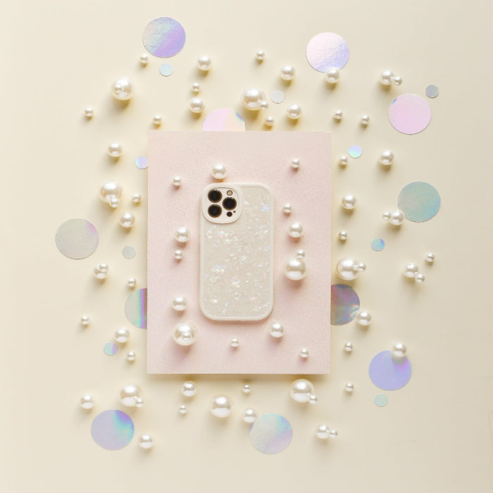 A smartphone in the Moonstone Pearl Phone Case by Coconut Lane, featuring a sparkly, translucent design, is placed on a light pink rectangular paper. Surrounding it are scattered white pearls and iridescent circular confetti on a light beige background, creating a decorative and elegant arrangement.