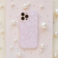 A smartphone encased in a Moonstone Pink Phone Case by Coconut Lane, glossy and textured, rests on a surface adorned with various sizes of pearls. The camera lenses are visible at the top left of the phone. The background features a slightly glittery pastel finish, adding an elegant touch.