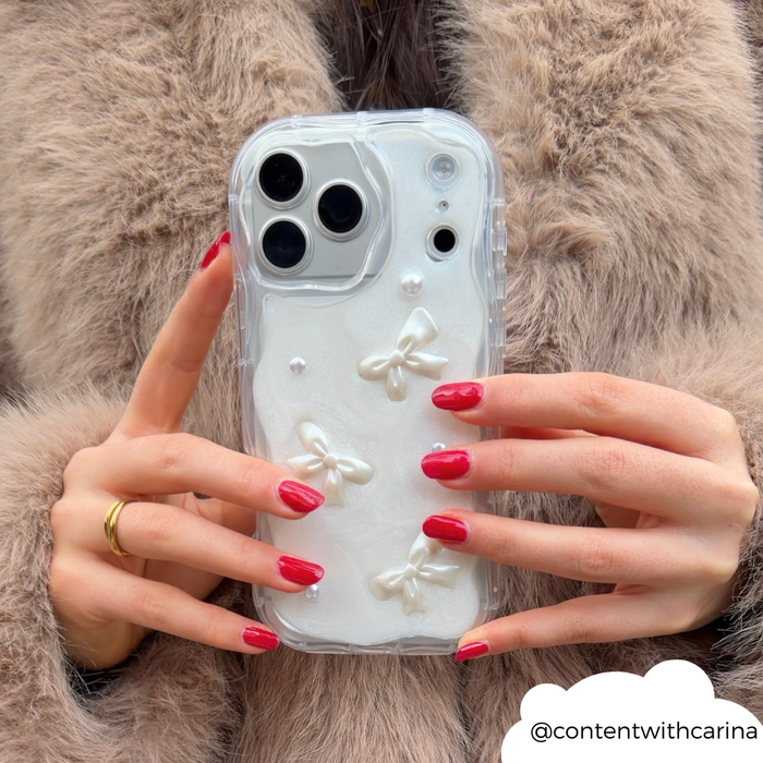 Red-nailed hand holds Coconut Lane Pearls & Bows phone case with pearl and bow details; fur coat in background.