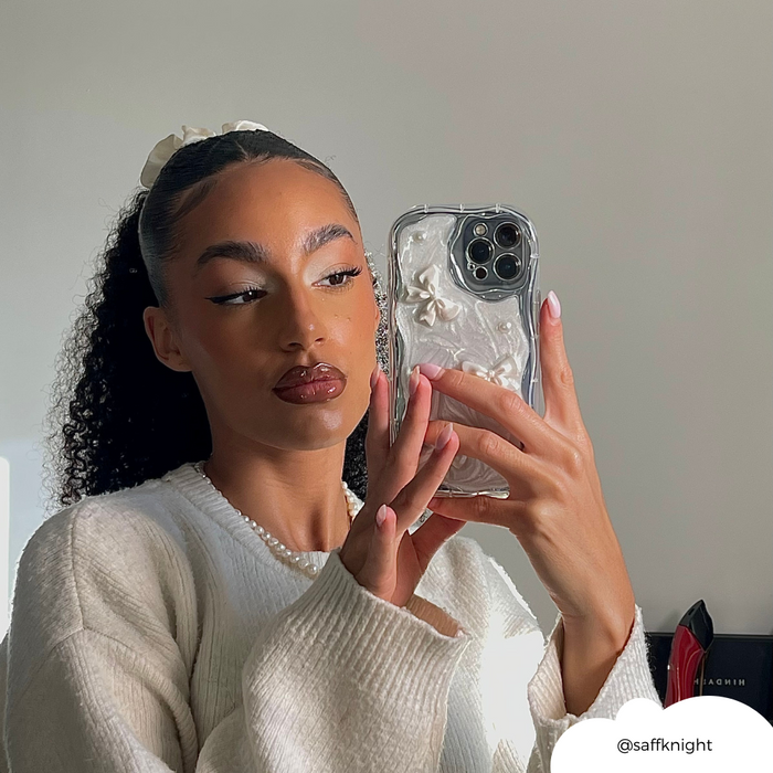 Woman with curly hair in a white sweater holds a phone with a pink Pearls & Bows case, taking a mirror selfie.