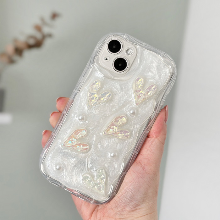 A hand holds an iPhone15 with an extra Coconut Lane Pearly Hearts Phone Case - Clear. The case showcases a pearlescent white design with raised heart shapes and small pearl-like embellishments. The phone is held in front of a blurred background.