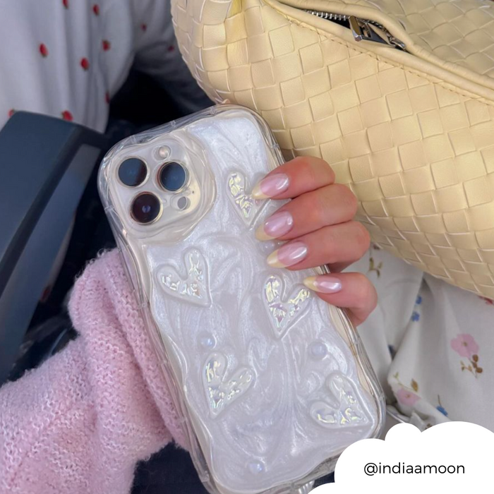 Hand with pastel French nails holds a clear phone case with pearly heart design next to a woven cream bag.