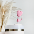 A white tumbler with a translucent straw is topped with Coconut Lane's Pink Heart Straw Cover, highlighting its flexible design. In the background, blurred pampas grass adds a touch of natural decor to the bright and minimalistic scene.