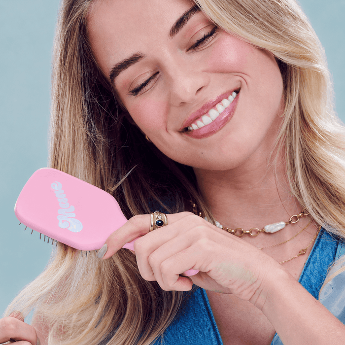 A smiling blonde woman with her eyes closed holds a pink hairbrush against her hair. She is wearing gold jewelry and a blue denim top, set against a solid light blue background.