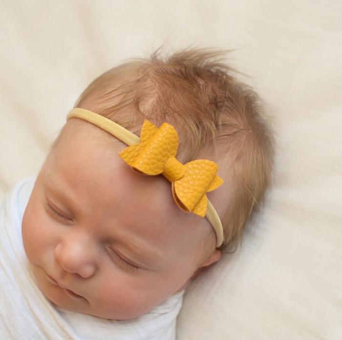 Newborn baby sleeping, wearing a mustard coloured leather bow headband