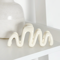 Cream Coconut Lane Squiggle Claw Clip on a shelf with a glossy white ceramic sculpture and a matte vase.