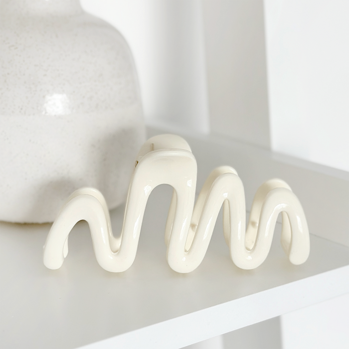 Cream Coconut Lane Squiggle Claw Clip on a shelf with a glossy white ceramic sculpture and a matte vase.