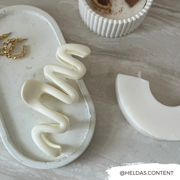 Cream squiggle claw clip on a stone tray with gold hoop earrings, white arc ornament, and ribbed candle holder.