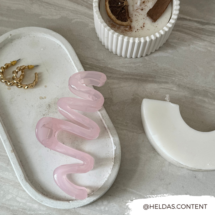 Pink squiggle claw clip on a tray with gold earrings, next to a candle and white decor on a gray surface.