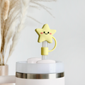 The Star Straw Cover by Coconut Lane is an adorable yellow, star-shaped silicone decoration with a cute smile and rosy cheeks. It is attached to a white and silver cylindrical object and photographed in front of a blurred background adorned with dried pampas grass.