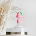 A close-up of a cup with a straw featuring Coconut Lane's Strawberry Straw Cover, a decorative pink strawberry-shaped silicone topper. The background is blurred, showing dry pampas grass against a light-colored wall.