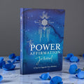 Blue journal titled 'The Power Affirmation Journal' with decorative elements on a gray background.
