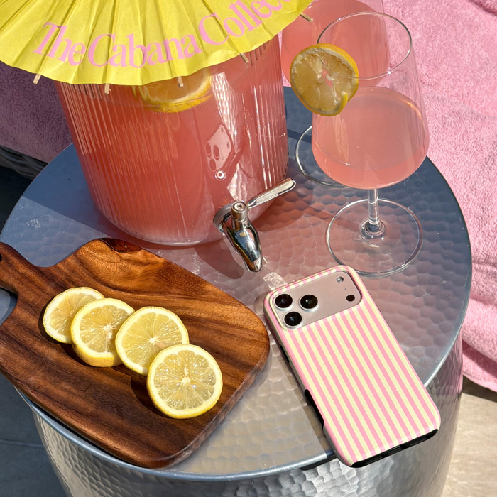 Pink lemonade with a wooden cutting board, sliced lemons, and a tough phone case with yellow and pink stripes on a glass table.