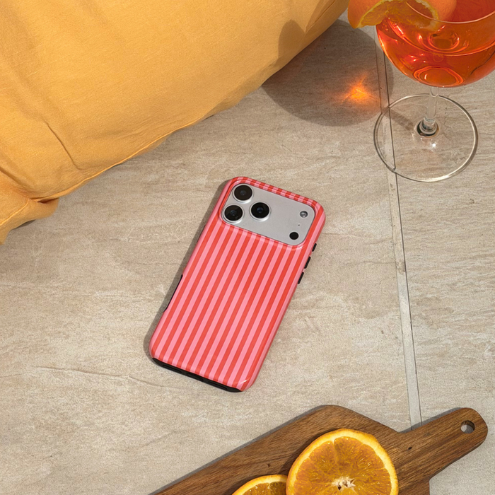 Phone with a pink and orange striped case on a tiled floor next to a glass of orange liquid and a cutting board with oranges.