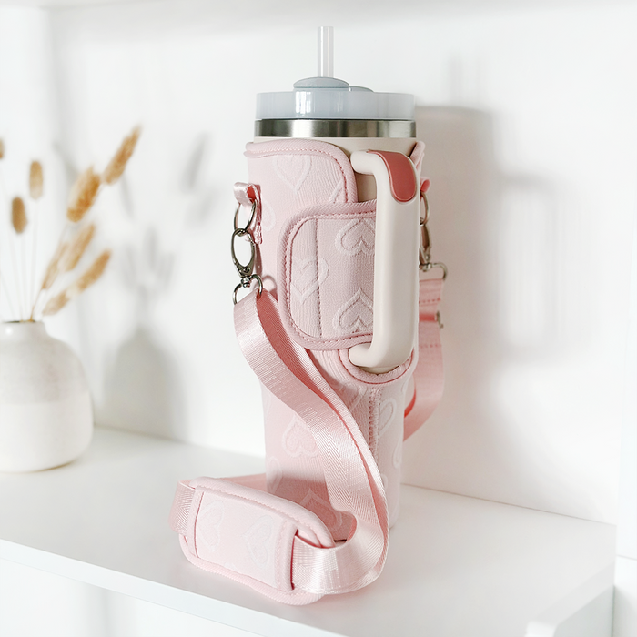 A Coconut Lane Tumbler Carry Case - Baby Pink Heart with a long strap securely holds a travel mug with a straw. The holder, adorned with handle and heart patterns, is placed on a white shelf next to a small vase containing dried wheat stalks, ensuring stylish tumbler protection.