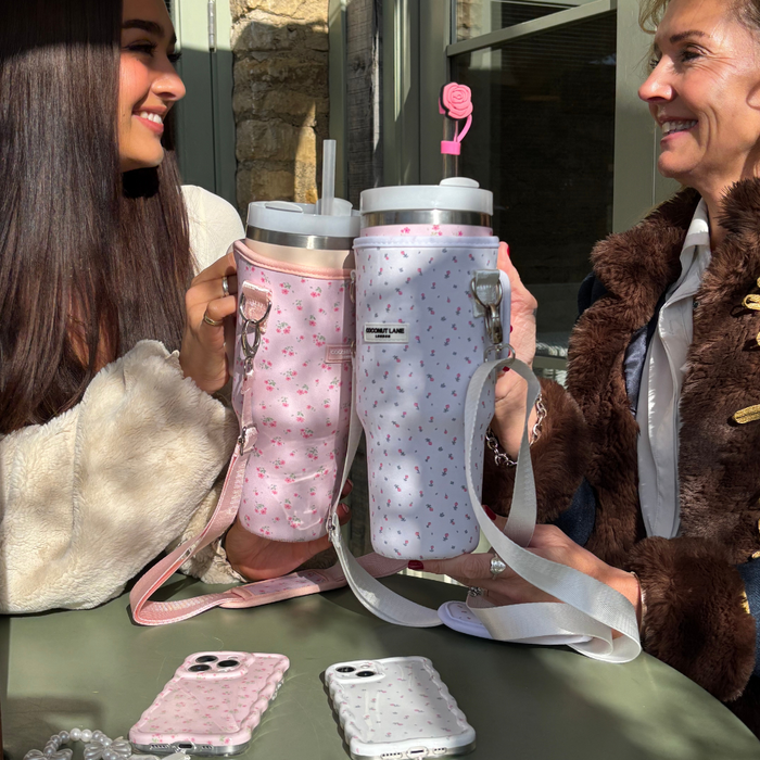 Two women smile at a table, each holding a Stanley Quencher in a pink floral Coconut Lane tumbler carry case.