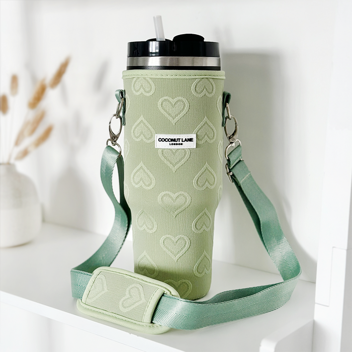 A green insulated Stanley Quencher tumbler, featuring a black lid and white straw, is showcased alongside a matching "Tumbler Carry Case - Sage Heart." The travel mug bag from Coconut Lane is decorated with heart patterns and includes a shoulder strap with the "Coconut Lane" label. In the background, dried plants and a white vase are visible.