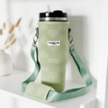 A green insulated Stanley Quencher tumbler, featuring a black lid and white straw, is showcased alongside a matching "Tumbler Carry Case - Sage Heart." The travel mug bag from Coconut Lane is decorated with heart patterns and includes a shoulder strap with the "Coconut Lane" label. In the background, dried plants and a white vase are visible.