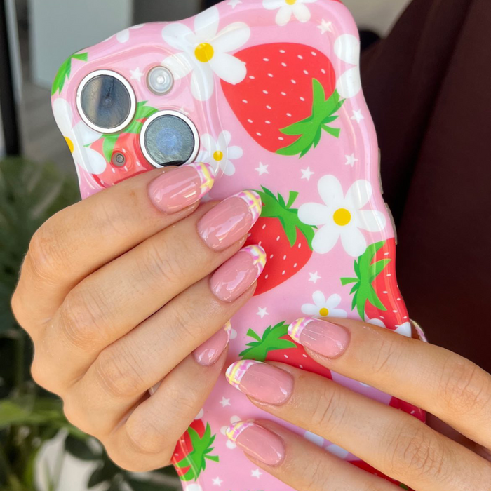 Pink-nailed hands hold a wavy phone case with strawberry and daisy pattern on a pink background.