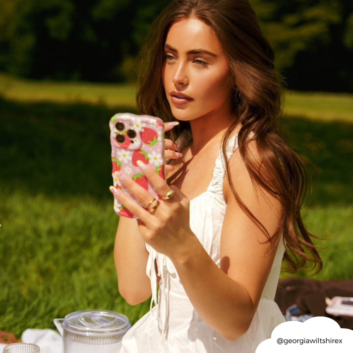 Woman in a white dress takes a selfie outdoors, holding a pink wavy phone case with red strawberry design.