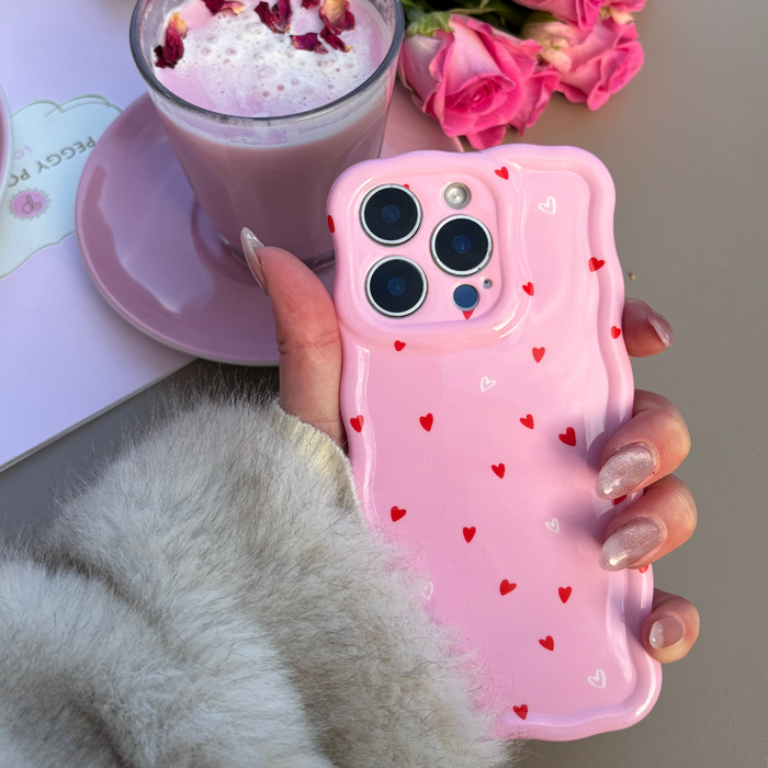 Hand in fur sleeve holds Coconut Lane wavy phone case with pink heart pattern, near a pink drink and pink roses.