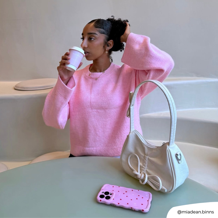 Woman in pink sweater drinks beside a Coconut Lane wavy phone case with cute heart pattern on a green table.