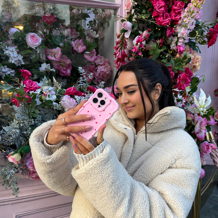 Woman in white fleece jacket holds pink wavy phone case with heart pattern, standing by a floral window.