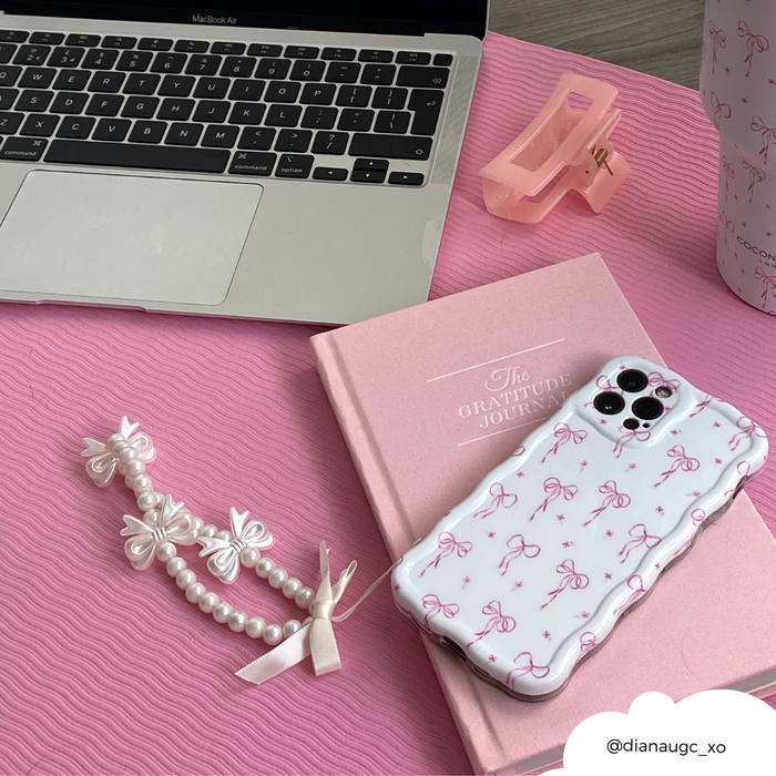Laptop, pink journal, wavy phone case with dainty bows, pearl accessory, and hair clip on a pink desk.