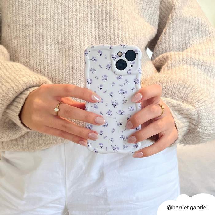 Woman in a beige sweater holding a periwinkle wavy phone case with a ditsy floral pattern and purple flowers.